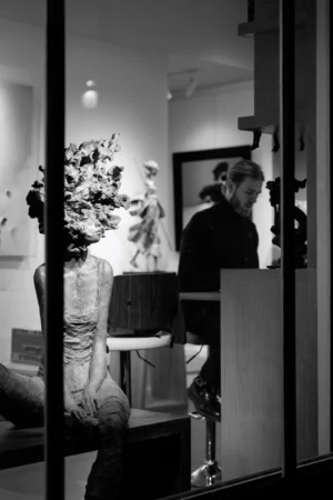 Philippe · 2001 x 3000 Cette photographie en noir et blanc capture une scène intérieure à travers une porte entrouverte. Le sujet principal semble être une femme assise, partiellement visible, avec un homme derrière elle. La composition met en valeur un espace minimaliste, avec des éléments artistiques (sculptures, cadre au mur) et une décoration sobre.Le cadrage est serré, créant une ambiance intimiste. La lumière douce et tamisée accentue les contrastes, ajoutant une touche dramatique. Les couleurs dominantes, bien que réduites au noir et blanc, suggèrent des tons clairs et sombres, renforçant l'atmosphère contemplative et élégante. Le style évoque une esthétique artistique et moderne.