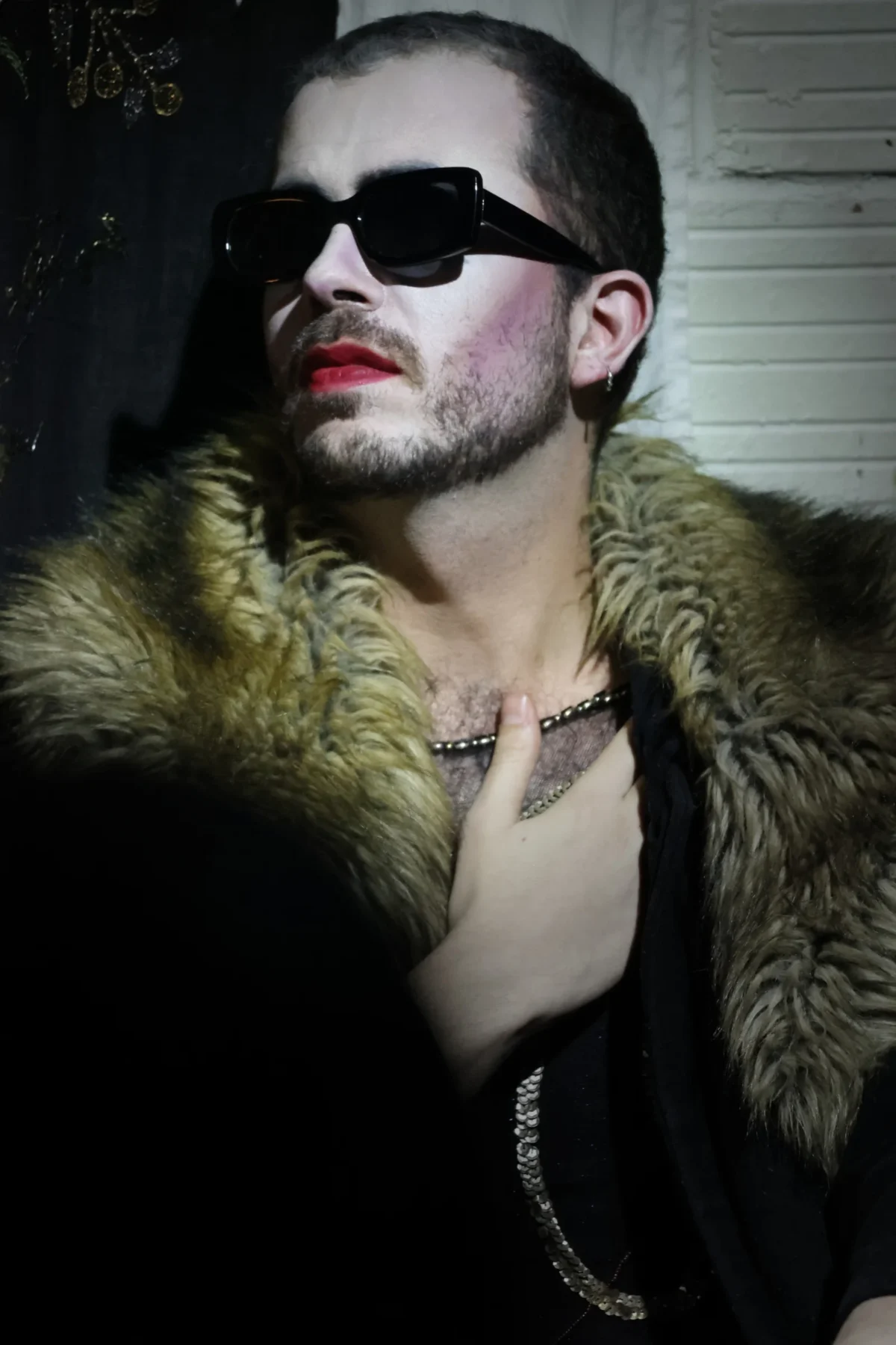 Olivier · 20/12/2024 · 3648 x 5472 Cette photographie met en scène un homme comme sujet principal, vêtu d'un long manteau en fourrure et portant des lunettes de soleil ainsi qu'un collier. Le cadrage est serré, centré sur son visage et sa poitrine.La lumière est tamisée, créant des ombres marquées et une ambiance mystérieuse. Les couleurs dominantes sont les tons sombres (noir, gris) avec des touches de vert et de rouge (lèvres, fourrure).Le style évoque un mélange de glamour et de provocation, renforcé par le maquillage rouge à lèvres. L'ambiance générale est à la fois sensuelle et énigmatique.