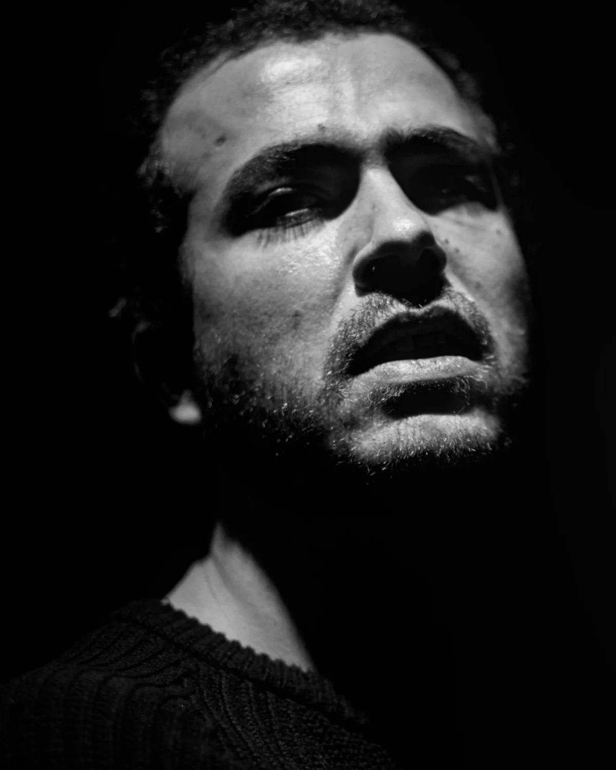 Olivier · 14/02/2025 · 2936 x 3670 Cette photographie met en scène un homme comme sujet principal, cadrée en gros plan. Son visage, légèrement incliné vers le haut, exprime une intensité mélancolique ou déterminée. La composition est épurée, centrée sur son regard et son expression.La lumière, contrastée et dramatique, crée des ombres marquées, accentuant les traits du visage. Les couleurs dominantes sont les noirs et gris, typiques d’un style noir et blanc.L’ambiance est sombre et théâtrale, évoquant une émotion profonde et une atmosphère intense. Le style rappelle le cinéma ou la photographie artistique classique.