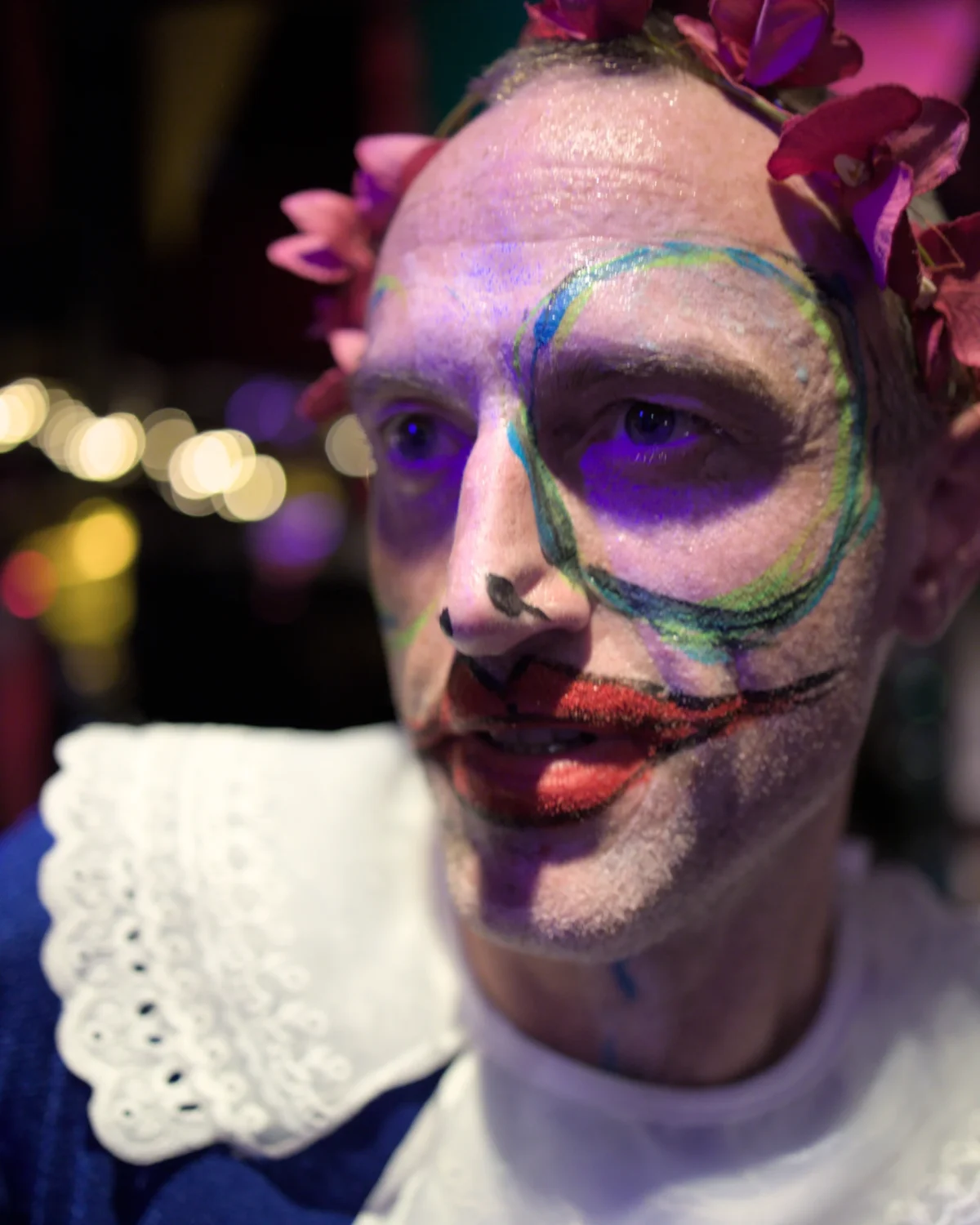 Olivier · 09/03/2025 · 3669 x 4587 Cette photographie met en scène un individu maquillé de manière théâtrale, évoquant un personnage clownesque ou grotesque. Le sujet principal est centré, avec un cadrage serré sur son visage.La lumière, douce et légèrement tamisée, met en valeur les détails du maquillage coloré (vert, bleu, rouge). Les couleurs dominantes sont vives et contrastées, créant une ambiance à la fois mystérieuse et festive. Le style rappelle le théâtre ou le carnaval, renforcé par les éléments décoratifs comme la fleur sur la tête. L'atmosphère est à la fois étrange et captivante.