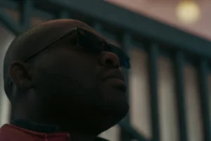 Don · 7008 x 4672 Cette photographie met en scène un homme comme sujet principal, cadré en gros plan. Il porte des lunettes de soleil et un vêtement rouge, avec une expression neutre ou légèrement concentrée. La composition est simple, axée sur son visage.La lumière semble douce mais contrastée, avec des ombres marquées sous ses yeux et sur son visage. Les couleurs dominantes sont les tons chauds (rouge, beige) et des teintes neutres. L’ambiance est sobre et légèrement dramatique, évoquant une atmosphère introspective ou cinématographique. Le style rappelle un plan typique de film ou de série.
