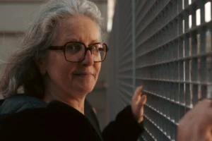 Don · 7008 x 4672 Cette photographie met en scène une femme d'âge mûr, sujet principal, portant des lunettes et un regard pensif. Le cadrage est serré sur son visage et sa main touchant une grille métallique.La composition est simple, axée sur les expressions et détails du visage. La lumière est naturelle et douce, éclairant principalement la femme par la gauche, créant des ombres légères. Les couleurs dominantes sont des tons neutres : gris, noir et beige, avec des touches de vert sur ses vêtements.L’ambiance est sobre et introspective, évoquant une atmosphère à la fois réaliste et légèrement mélancolique.
