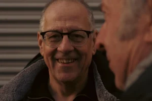 Don · 7008 x 4672 Cette photographie met en scène un homme souriant comme sujet principal, partiellement cadré en gros plan. Son visage est éclairé, avec des lunettes et une barbe grisonnante. La composition inclut une partie d’un autre individu en arrière-plan, légèrement flou.Le cadrage est serré, centré sur le visage principal. La lumière est douce et naturelle, créant des ombres subtiles. Les couleurs dominantes sont des tons neutres : gris, beige et noir, avec des touches de bleu et de gris clair sur les vêtements.L’ambiance est chaleureuse et conviviale, évoquant une scène intime et détendue. Le style est simple et réaliste.