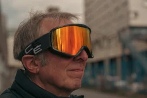 Don · 7008 x 4672 Cette photographie met en scène un homme d'âge mûr comme sujet principal, portant des lunettes de ski aux verres teintés orange. Le cadrage est serré sur son visage, avec un arrière-plan flou montrant des bâtiments et un ciel dégagé. La lumière est naturelle, probablement en journée, avec un éclairage doux et homogène. Les couleurs dominantes sont les tons froids (bleu des bâtiments) et chauds (orange des verres, teint de peau). Le style est réaliste et spontané, créant une ambiance à la fois sportive et détendue, évoquant une activité hivernale en milieu urbain.