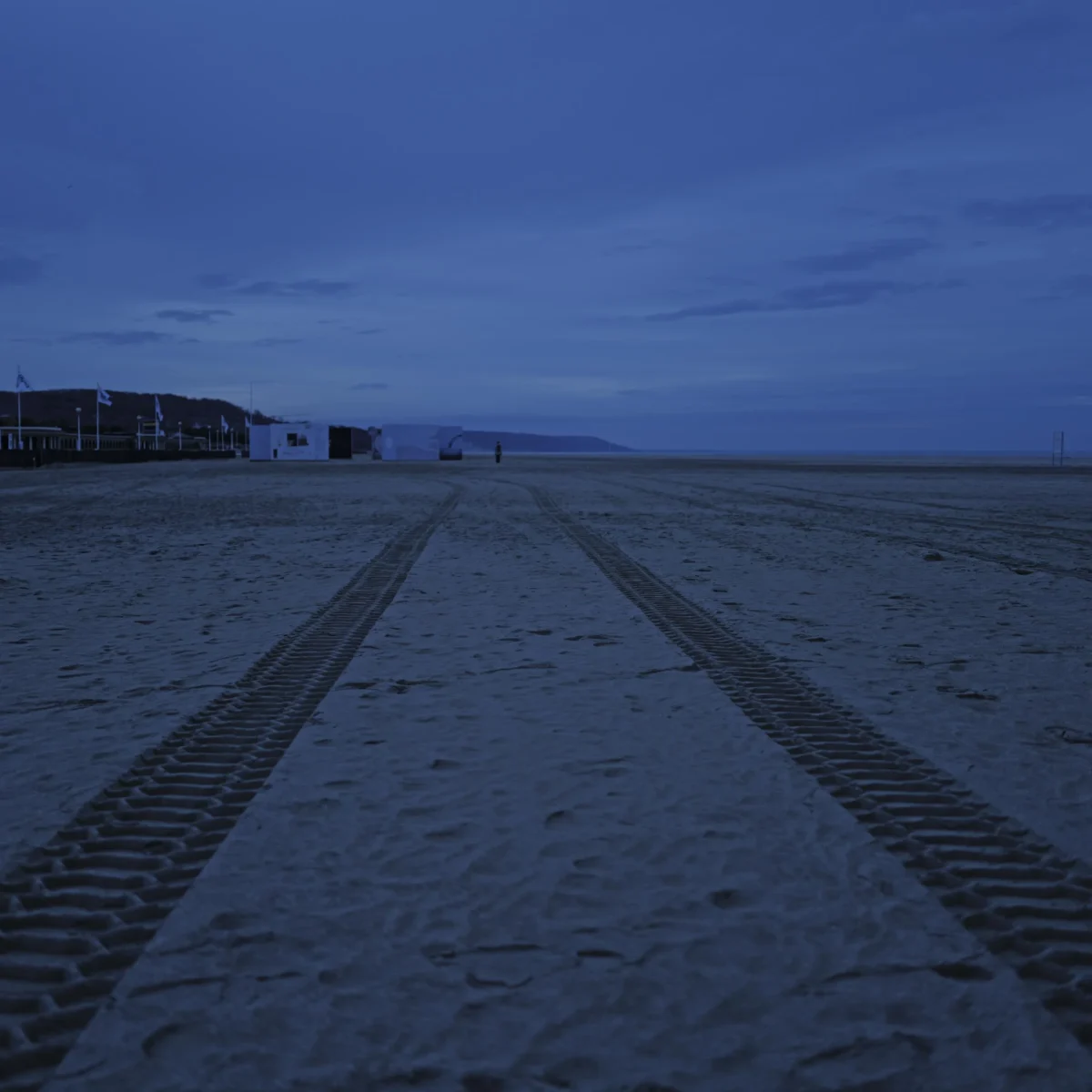 Jean-Paul · 12/12/2025 · 3000 x 3000 Cette photographie capture une plage déserte avec des traces de pas ou de véhicules tout-terrain dans le sable. Le sujet principal est la perspective des sillons menant vers l’horizon, où l’on distingue une structure urbaine à gauche.La composition repose sur des lignes directrices convergentes, renforçant la profondeur. Le cadrage est large, englobant une vue panoramique. La lumière, tamisée, crée une ambiance crépusculaire ou nocturne. Les couleurs dominantes sont les bleus profonds et les gris, avec des touches de blanc dans le sable.Le style est épuré et contemplatif, évoquant une atmosphère calme et mystérieuse.