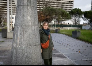Muriel · 1080 x 773 Cette photographie met en scène une femme comme sujet principal, debout près d'une colonne en pierre. Elle porte une veste verte et un sac rouge, ajoutant une touche de couleur vive.La composition est simple et équilibrée, avec un cadrage serré sur la femme et un arrière-plan urbain. La lumière semble naturelle, légèrement diffuse, créant des ombres douces. Les couleurs dominantes sont les tons neutres (gris de la pierre et du sol) contrastés par le rouge du sac et le vert de sa veste.Le style est réaliste et documentaire, évoquant une ambiance calme et urbaine.