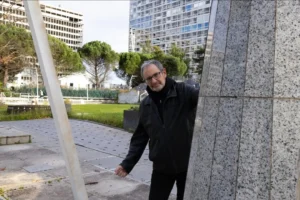 Muriel · 1080 x 720 Cette photographie met en scène un homme souriant, sujet principal, posé contre une colonne en pierre grise. La composition est simple et naturelle, avec un cadrage serré sur le sujet et un arrière-plan urbain.La lumière semble naturelle, probablement en journée, avec des tons doux. Les couleurs dominantes sont les gris (bâtiments, colonne), les verts (arbres, pelouse) et les teintes sombres du manteau de l’homme. Le style est réaliste et documentaire, capturant un moment spontané. L’ambiance est calme et urbaine, avec une touche de convivialité grâce au sourire du sujet.