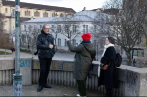 Muriel · 1080 x 717 Cette photographie montre trois personnes en extérieur, près d’un pont. Le sujet principal semble être une interaction entre elles, avec une personne en rouge marquant l’attention.La composition est dynamique, avec un cadrage serré sur les individus. L’arrière-plan inclut des bâtiments et des arbres dénudés. La lumière est naturelle, un peu grise, typique d’un jour d’hiver. Les couleurs dominantes sont les tons froids (gris, bleu) et quelques touches chaudes (vêtements rouges et noirs).Le style est documentaire et réaliste, capturant une scène du quotidien. L’ambiance est calme et un peu mélancolique.