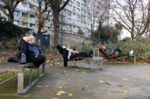 Muriel · 1080 x 716 Cette photographie montre des mannequins habillés en costumes d'affaires, disposés comme des personnes endormies ou inconscientes sur des bancs publics.La composition met en scène une ambiance surréaliste et inquiétante. Le cadrage inclut un parc urbain avec des bâtiments en arrière-plan. La lumière est naturelle, probablement en journée, avec des ombres marquées. Les couleurs dominantes sont les tons froids et neutres, avec des touches de gris, bleu et vert. Le style est réaliste, mais l'ambiance est troublante, évoquant une critique sociale ou une allégorie de l'épuisement moderne.