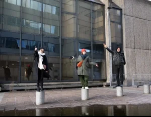Muriel · 1080 x 836 Cette photographie montre trois personnes en mouvement devant un immeuble moderne. Le sujet principal semble être une scène dynamique, évoquant une performance artistique ou une danse improvisée.La composition est symétrique, avec les trois individus alignés verticalement. Le cadrage est serré sur les sujets, avec un arrière-plan architectural reflétant la lumière. Les couleurs dominantes sont neutres (gris, beige) avec des touches de rouge et noir sur les vêtements.Le style est réaliste et spontané, capturant un moment vif. L’ambiance est à la fois urbaine et artistique, avec une lumière naturelle qui accentue les contours et les reflets.