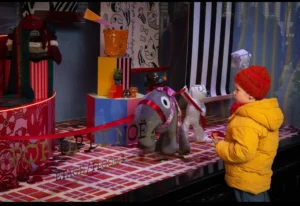 Cette photographie montre une scène de film ou de série animée. Le sujet principal est un enfant en manteau jaune observant un éléphant anthropomorphe en costume de fête, entouré de décorations festives.La composition est dynamique, avec un cadrage serré sur les personnages et les éléments décoratifs. Les couleurs dominantes sont vives : rouge, jaune, bleu et blanc. La lumière semble douce et uniforme, créant une ambiance chaleureuse et ludique. Le style est celui d’une animation colorée, typique des productions familiales, évoquant la joie et la magie des fêtes.