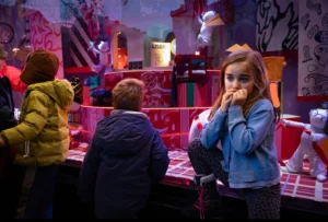 Cette photographie montre une jeune fille comme sujet principal, tenant un téléphone à l’oreille, dans un lieu animé et coloré. La composition met en avant un groupe de personnes en arrière-plan, légèrement floues, créant une profondeur.Le cadrage est serré sur la jeune fille, avec une lumière vive et contrastée. Les couleurs dominantes sont vives et chaudes : rouges, roses et teintes pastel. L’ambiance est dynamique et festive, évoquant un espace ludique ou une fête foraine. Le style est réaliste et spontané, capturant un moment du quotidien.