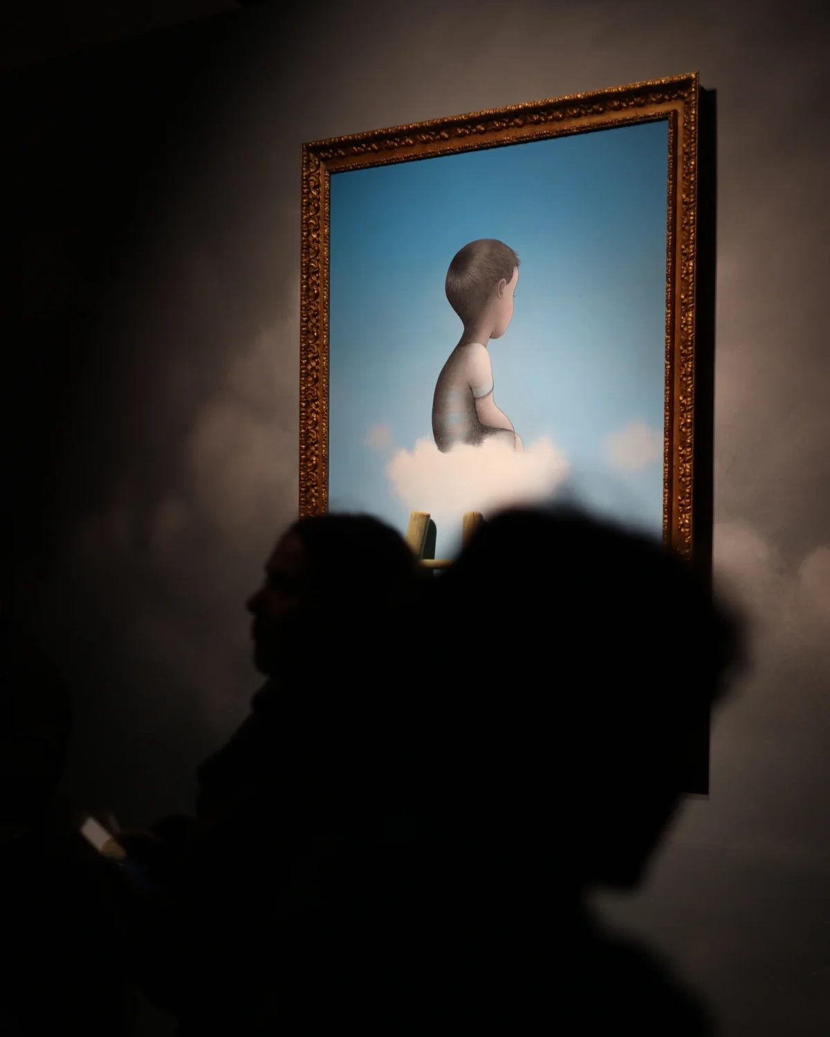 Olivier · 29/11/2024 · 3647 x 4559 Cette photographie met en scène un tableau surréaliste accroché au mur, représentant une figure féminine émergeant d’un nuage. Le sujet principal est ce tableau, encadré par une bordure dorée.La composition joue sur le contraste entre la lumière douce éclairant la peinture et l’ombre des silhouettes en contre-jour. Le cadrage, serré, isole le tableau et les deux personnes dont les visages sont dans l’obscurité, créant une ambiance mystérieuse.Les couleurs dominantes sont les bleus froids du ciel et les tons chauds du cadre. Le style évoque le surréalisme, renforcé par l’éclairage dramatique et l’atmosphère contemplative.