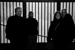 Tery · 06/12/2025 · 6720 x 4480 Cette photographie en noir et blanc montre quatre personnes posant ensemble devant un fond strié vertical. Le sujet principal est le groupe, aligné de manière informelle.Le cadrage est serré, mettant en valeur les visages et les épaules. La lumière est douce et uniforme, créant des contrastes modérés. Les couleurs dominantes sont les nuances de gris, typiques du noir et blanc.Le style est sobre et documentaire, évoquant une ambiance sérieuse et intime. L'image dégage une impression de simplicité et de sincérité.