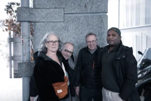 Tery · 06/12/2025 · 6720 x 4480 Cette photographie montre quatre personnes posant devant une structure architecturale moderne. Le sujet principal semble être le groupe, avec une femme au premier plan tenant un sac en bandoulière.La composition est symétrique et serrée, avec un cadrage en plan moyen. La lumière est naturelle, douce mais un peu froide, créant des contrastes modérés. Les couleurs dominantes sont neutres, avec des teintes grises et noires, adoucies par quelques touches de brun et de beige.Le style est sobre et documentaire, évoquant une ambiance professionnelle ou informelle, sobre et contemporaine.