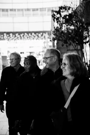 Tery · 06/12/2025 · 4480 x 6720 Cette photographie en noir et blanc montre quatre personnes marchant côte à côte, formant le sujet principal. La composition est simple et linéaire, avec un cadrage serré sur les individus.La lumière est douce, créant des contrastes subtils entre les visages et l’arrière-plan. Les ombres et les reflets ajoutent une touche dramatique.Les couleurs dominantes sont les nuances de gris, typiques du noir et blanc, renforçant une ambiance sobre et intemporelle.Le style est documentaire et naturel, évoquant une scène du quotidien. L’ambiance est calme et contemplative, presque mélancolique.