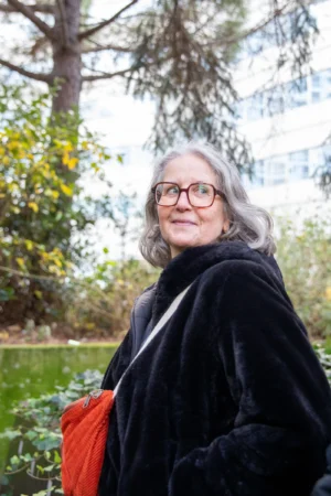 Tery · 3809 x 5714 Cette photographie met en scène une personne âgée comme sujet principal, souriante et détendue. Le cadrage est serré, centré sur elle, avec un arrière-plan flou de végétation (arbres et buissons).La lumière naturelle, douce et légèrement tamisée, crée une ambiance chaleureuse et apaisante. Les couleurs dominantes sont les tons naturels : vert des feuilles, gris-bleu du manteau, et rouge vif du sac. Le style est simple et naturel, évoquant une ambiance authentique et paisible, presque intimiste.