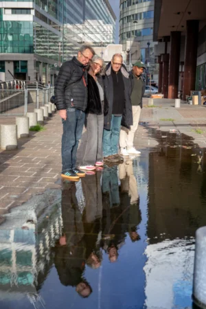 Tery · 4301 x 6451 Cette photographie montre trois personnes debout sur un trottoir urbain, reflétées dans une flaque d’eau. Le sujet principal est leur interaction joyeuse, avec un cadrage serré sur eux.La composition utilise la symétrie des reflets pour créer un effet visuel intéressant. La lumière semble naturelle, probablement en journée, avec des tons clairs. Les couleurs dominantes sont les gris urbains, les teintes sombres des vêtements et les reflets bleutés de l’eau.Le style est documentaire et spontané, évoquant une ambiance détendue et presque ludique dans un cadre urbain moderne.