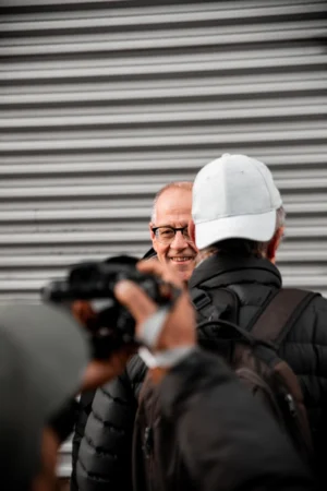 Tery · 4480 x 6720 Cette photographie met en scène un homme comme sujet principal, portant une casquette blanche et un sac à dos, occupé à prendre une photo avec un appareil reflex. Le cadrage est serré, centré sur lui, avec un arrière-plan flou composé de bandes horizontales métalliques.La lumière semble naturelle et douce, créant des ombres peu marquées. Les couleurs dominantes sont neutres, avec des tons gris et noirs, adoucis par des touches de blanc et de beige. Le style est documentaire et spontané, évoquant une ambiance authentique et professionnelle, typique d’un photographe en action.