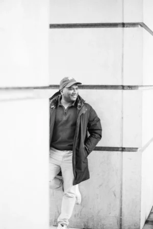Tery · 4480 x 6720 Cette photographie en noir et blanc met en scène un homme comme sujet principal, vêtu d’un manteau long, d’un chapeau et de bottes, posant contre un mur strié. Le cadrage est serré, centré sur lui, avec un arrière-plan minimaliste.La lumière douce et naturelle crée des contrastes subtils, accentuant les textures. L’ambiance est sobre et introspective, évoquant une touche vintage ou documentaire. Les tons clairs et foncés renforcent le côté mélancolique et authentique de la scène, typique d’un style photographique classique et épuré.