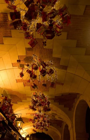 Cette photographie met en scène des **bouquets suspendus** décorant un plafond voûté. Le **sujet principal** est ces arrangements floraux, mêlant fleurs séchées et éléments végétaux.La **composition** est verticale, avec un **cadrage serré** sur les bouquets, accentuant leur élégance. La **lumière** douce et tamisée crée une ambiance chaleureuse et mystérieuse. Les **couleurs dominantes** sont des tons **beige, dorés et ocres**, rehaussés par des touches de rouge et de vert.Le **style** évoque une atmosphère **romantique et vintage**, renforcée par les structures architecturales anciennes. L’**ambiance** est à la fois **intime et solennelle**, évoquant une décoration soignée et raffinée.