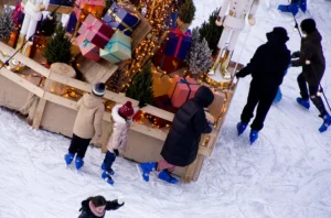Cette photographie montre un groupe de personnes patinant sur une patinoire en plein air. Le sujet principal est une famille ou des amis en train de glisser ensemble.La composition est dynamique, avec un cadrage aérien offrant une vue d'ensemble. Les couleurs dominantes sont les tons froids : blanc de la glace et de la neige, bleu du ciel, et quelques touches chaudes avec les vêtements des patineurs.La lumière semble naturelle, probablement en journée. L’ambiance est festive et joyeuse, renforcée par les décorations lumineuses et les couleurs vives des accessoires. Le style est documentaire et captivant.