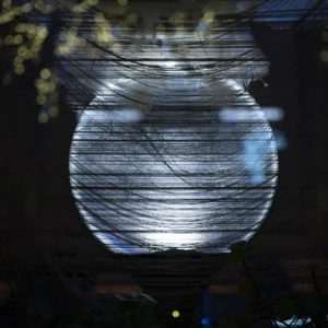 Cette photographie met en scène une **sphère lumineuse** suspendue, probablement une installation artistique ou un objet décoratif. Le **sujet principal** est cette boule translucide, traversée par des **lignes lumineuses** internes, créant un effet de mouvement et de profondeur.Le **cadrage** est serré, centré sur la sphère, avec un **flou d’arrière-plan** qui isole le sujet. La **lumière** est douce mais intense, avec des reflets bleutés et des éclats jaunes en périphérie. Les **couleurs dominantes** sont des teintes froides (bleu, gris) et des touches chaudes (jaune).Le style évoque une **ambiance onirique et futuriste**, presque hypnotique, renforcée par les jeux de lumière et les traînées lumineuses.
