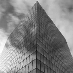 Cette photographie met en scène un **gratte-ciel moderne** comme sujet principal, cadré en **plan serré** depuis sa base, accentuant sa hauteur et ses lignes géométriques.La **composition** est épurée, centrée sur l’architecture. La **lumière**, douce et diffuse, crée des contrastes subtils entre les ombres et les reflets des vitres. Les **couleurs dominantes** sont en **niveaux de gris**, renforçant un style **minimaliste et abstrait**.L’ambiance est **sobre et mystérieuse**, évoquant puissance et modernité, avec une touche dramatique due aux nuages en arrière-plan.