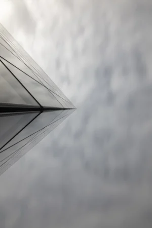 Cette photographie met en scène un **angle d’un toit ou d’une structure métallique** en perspective convergente. La **composition** repose sur des lignes géométriques qui guident le regard vers l’horizon.Le **cadrage** serré et l’**angle bas** accentuent la profondeur. La **lumière**, douce et diffuse, crée des contrastes subtils. Les **couleurs dominantes** sont des teintes grises et bleutées, renforçant une ambiance **minérale et épurée**.Le style, **abstrait et minimaliste**, évoque une sensation de calme et de modernité, avec une **ambiance presque onirique**.