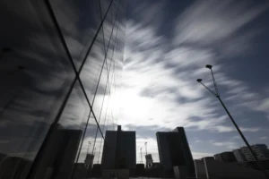 Cette photographie met en scène un **ciel nuageux** traversé par des **structures verticales** (poteaux ou câbles), encadrant le soleil en arrière-plan. Le **sujet principal** est la lumière filtrant à travers les nuages, créant un effet dramatique.La **composition** joue sur les lignes verticales et horizontales, avec un **cadrage serré** sur les éléments urbains en bas. La **lumière** est douce mais contrastée, avec des ombres marquées. Les **couleurs dominantes** sont les gris et noirs, adoucis par les teintes claires du soleil.Le **style** est épuré et minimaliste, évoquant une **ambiance contemplative et presque mystique**.