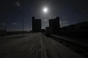 Cette photographie capture une route déserte sous un éclairage nocturne. Le **sujet principal** est la voie presque vide, avec des **silhouettes de bâtiments** en arrière-plan. La **composition** repose sur une perspective frontale et symétrique, renforcée par les lignes droites de la route et les **lignes horizontales** des rails.Le **cadrage** est serré, créant un effet de profondeur. La **lumière**, douce et contrastée, provient probablement de la lune et d’un éclairage urbain, donnant une ambiance **sombre et mystérieuse**. Les **couleurs dominantes** sont les noirs et gris, avec des touches de bleu nuit. Le style est **minimaliste et dramatique**, évoquant une **ambiance solitaire et onirique**.