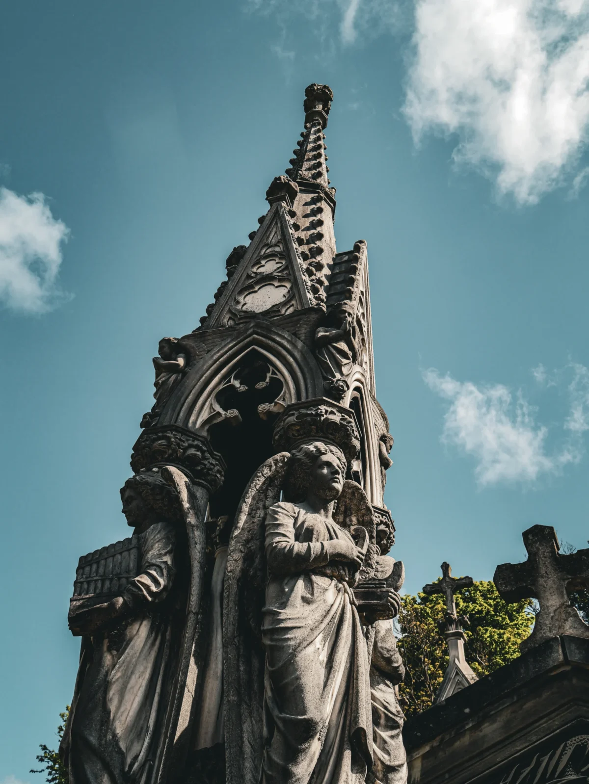 Laurent · 2160 x 2877 Cette photographie met en scène une **statue gothique** détaillée, probablement un ange ou une figure religieuse, intégrée à une structure architecturale ornée.La **composition** est verticale, avec un **cadrage serré** sur le haut de la sculpture, accentuant son élévation. La **lumière naturelle**, douce et contrastée, souligne les textures et les ombres, créant un effet dramatique. Les **couleurs dominantes** sont les gris et noirs de la pierre, rehaussés par le bleu clair du ciel.Le **style** est **gothique**, avec des motifs complexes et une ambiance **mystérieuse et solennelle**, renforcée par l'arrière-plan flou et les nuages.