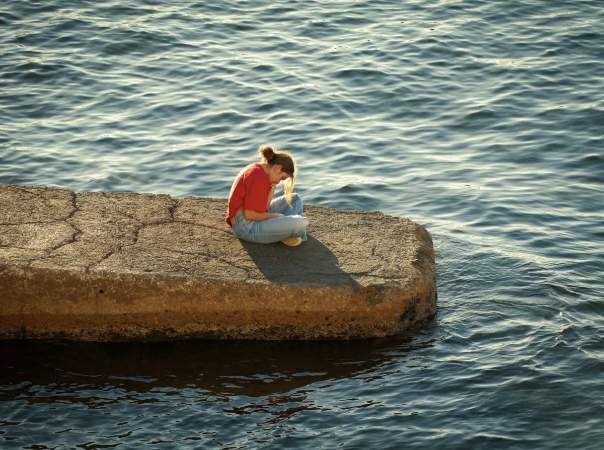Laurent · 2899 x 2160 Cette photographie met en scène une personne assise seule sur un bord de quai en béton, penchée en avant, les mains entre les genoux. La composition est épurée, centrée sur le sujet et l’eau calme en arrière-plan.Le cadrage serré isole le personnage, créant une ambiance introspective. La lumière, douce et naturelle, semble provenir de la gauche, adoucissant les contours. Les couleurs dominantes sont les tons gris du béton et de l’eau, contrastés par le rouge vif du vêtement du sujet.Le style est réaliste et sobre, évoquant une atmosphère mélancolique et contemplative.