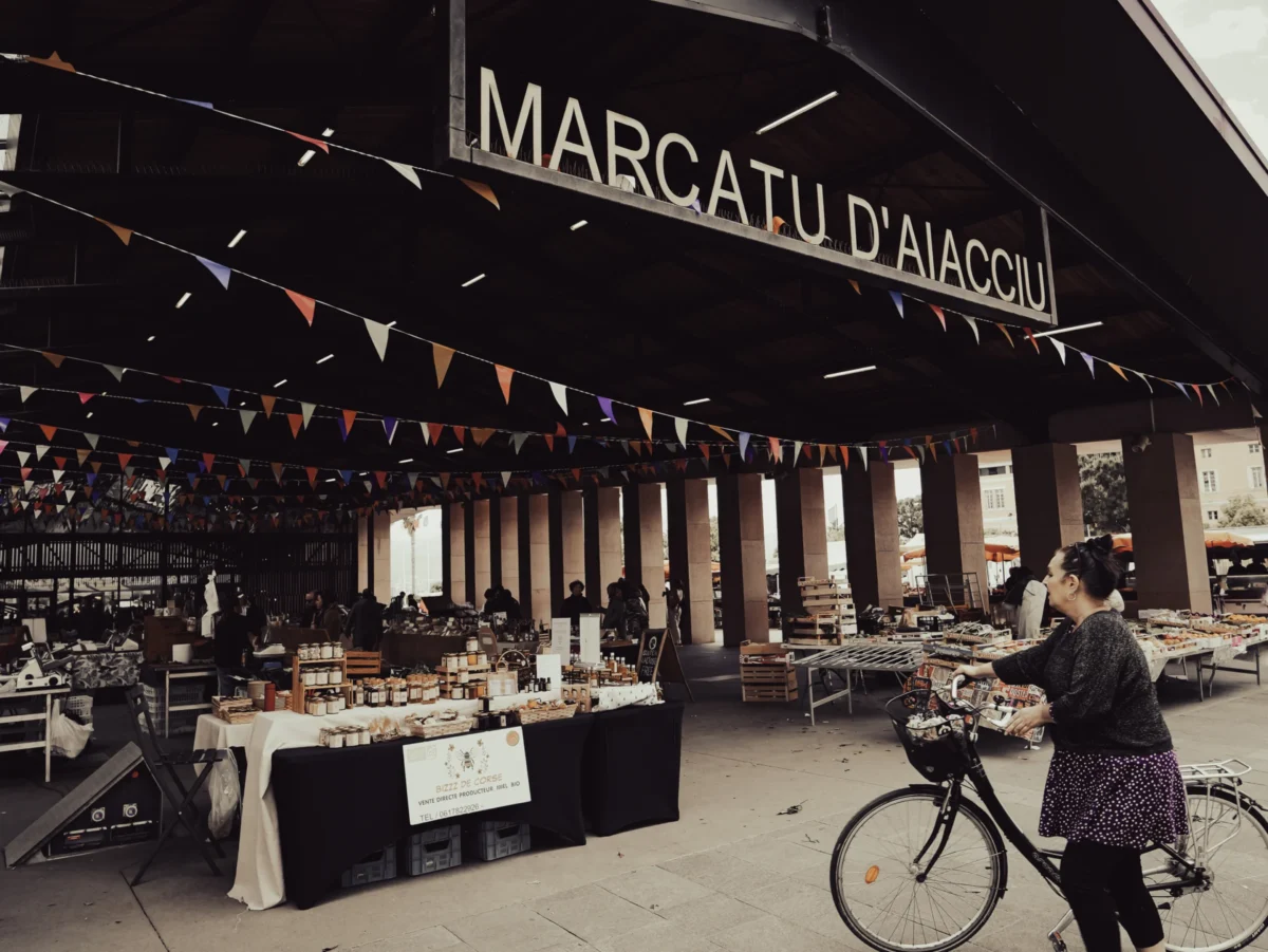 Laurent · 2877 x 2160 Cette photographie montre un marché couvert nommé **« Marcatu d’Aiacciu »**. Le sujet principal est une personne à vélo, en train de prendre une photo ou de filmer l’entrée du marché.La composition met en valeur les étals garnis de produits locaux sous une structure en bois. Le cadrage inclut des drapeaux suspendus et une ambiance animée. La lumière est naturelle, douce, avec des ombres légères, créant une atmosphère claire et accueillante. Les couleurs dominantes sont les tons chauds (bois, brun) et neutres, adoucis par des touches de vert et de blanc.Le style est documentaire et authentique, évoquant une ambiance artisanale et conviviale.