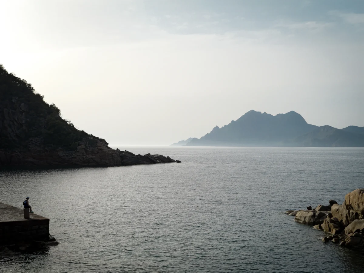 Laurent · 2877 x 2160 Cette photographie capture une scène côtière paisible. Le **sujet principal** est un **paysage marin** avec des montagnes en arrière-plan et une étendue d’eau calme.La **composition** est équilibrée, avec une ligne d’horizon centrale et des rochers en premier plan. Le **cadrage** est large, englobant une vaste perspective.La **lumière** est douce, probablement en fin de journée, créant une ambiance sereine. Les **couleurs dominantes** sont des teintes bleutées et grises, avec des nuances douces.Le **style** est réaliste et épuré, évoquant une **ambiance calme et contemplative**, presque mélancolique.