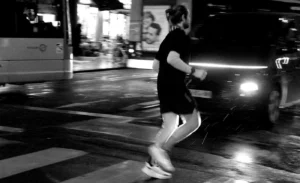 Franck T. · 20/12/2025 · 1400 x 854 Cette photographie en noir et blanc met en scène un homme en mouvement, probablement en train de courir ou de faire du sport. Le sujet principal est flou, suggérant un mouvement rapide, tandis que l’arrière-plan est légèrement net, révélant des véhicules et un environnement urbain nocturne.Le cadrage est dynamique, avec une composition qui capture l’action. La lumière est tamisée, avec des éclairages artificiels (voitures, enseignes) créant des contrastes marqués. Les couleurs dominantes, bien que monochromes, jouent sur les nuances de gris et de noir, renforçant l’ambiance nocturne et énergique. Le style est documentaire et spontané, évoquant une scène authentique et vivante.