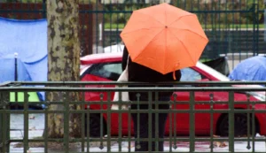 Franck T. · 20/12/2025 · 1400 x 808 Cette photographie montre une personne vue de dos, tenant un **parapluie orange** comme sujet principal. La composition met en avant un **cadrage serré**, avec des barrières métalliques en premier plan.Les couleurs dominantes sont les **tons froids (bleu, gris)** et l'**orange vif** du parapluie. La lumière semble naturelle, mais diffuse, créant une ambiance un peu sombre et humide.Le style est **documentaire et réaliste**, évoquant une scène urbaine quotidienne. L’ambiance est **calme et mélancolique**, renforcée par les couleurs contrastées et l’absence de visages.