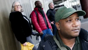 Franck T. · 06/12/2025 · 1400 x 795 Cette photographie met en avant un homme en premier plan, vêtu d’un chapeau vert et d’un pull sombre, avec un regard direct. En arrière-plan, plusieurs personnes attendent, habillées chaudement (veste rouge, noir, etc.).Le cadrage est serré sur l’homme, avec une profondeur de champ réduisant les détails des autres. La lumière semble naturelle, légèrement tamisée, créant des contrastes doux. Les couleurs dominantes sont les tons froids (gris, verts) et chauds (rouge, noir).Le style est documentaire, réaliste, capturant une scène urbaine quotidienne. L’ambiance est sobre et légèrement intimiste, évoquant une attente ou un moment de pause dans un lieu public.