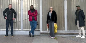 Franck T. · 06/12/2025 · 1400 x 703 Cette photographie montre quatre personnes debout devant une porte métallique. Le sujet principal semble être leur interaction ou leur attente.La composition est symétrique et linéaire, avec les individus alignés verticalement. Le cadrage est serré, centré sur les personnages. La lumière est naturelle et diffuse, sans ombres marquées.Les couleurs dominantes sont sobres : noir, gris, bleu et rouge. L’ambiance est sobre et urbaine, avec une touche de neutralité et de modernité. Le style est documentaire, capturant un moment du quotidien.