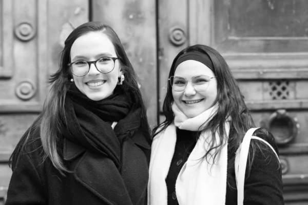 Claude · 5185 x 3457 Cette photographie en noir et blanc montre deux femmes souriantes posant côte à côte. Elles semblent détendues, avec des expressions amicales.La composition est symétrique, mettant en valeur les deux sujets principaux. Le cadrage est serré, centré sur elles, avec un arrière-plan flou composé de portes anciennes.La lumière est douce et naturelle, éclairant uniformément les visages. Les tons dominants sont les gris et noirs, typiques du noir et blanc, renforçant une ambiance intemporelle et chaleureuse. Le style est simple et spontané, évoquant une ambiance conviviale et authentique.