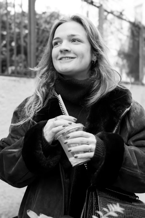 Claude · 4000 x 6000 Cette photographie en noir et blanc met en scène une femme comme sujet principal, souriante et détendue. Elle porte un manteau à motifs et tient un objet entre ses mains.Le cadrage est serré, centré sur son visage et sa poitrine, avec un arrière-plan flou montrant des éléments urbains. La lumière est douce et naturelle, éclairant son visage de manière homogène. L'absence de couleurs accentue les contrastes et les textures, comme les motifs du manteau.Le style est simple et spontané, évoquant une ambiance chaleureuse et intime, presque documentaire.