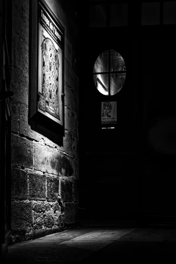 Claude · 4000 x 6000 Cette photographie en noir et blanc capture un couloir étroit et sombre. Le sujet principal est une porte ou un passage partiellement éclairé, avec des jeux d'ombres marquées. La composition repose sur un cadrage serré, mettant en valeur les textures des murs en pierre et les détails architecturaux, comme une plaque et un cadran circulaire.La lumière, provenant d'une source latérale, crée des contrastes forts et dramatise l'ambiance. Les couleurs dominantes sont les tons de gris, renforçant l'atmosphère mystérieuse et presque gothique. Le style est minimaliste et évocateur, évoquant une sensation de solitude et de mélancolie.
