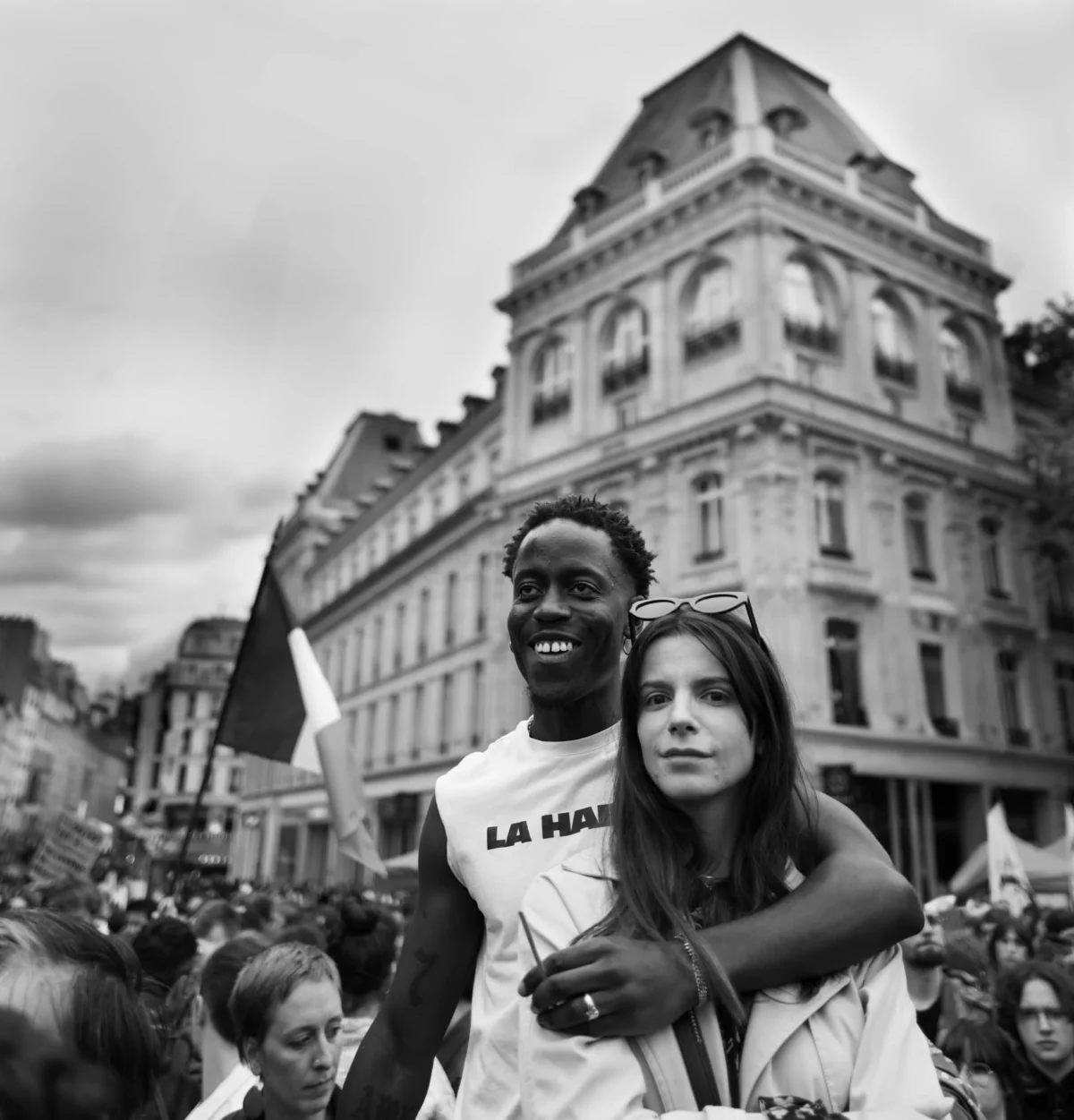 Pascal SC · 03/07/2024 · 1823 x 1900 Cette photographie en noir et blanc capture deux jeunes gens au premier plan, un homme et une femme, souriants et proches. Le sujet principal est leur complicité, mise en valeur par le cadrage serré.L’arrière-plan montre une foule et un bâtiment architectural classique, suggérant une manifestation ou rassemblement urbain. La lumière est douce, avec des contrastes modérés, accentuant les textures et les visages. L’ambiance est chaleureuse et intimiste malgré la foule, renforcée par le flou des éléments périphériques.Le style est documentaire et spontané, évoquant un moment authentique et émouvant.