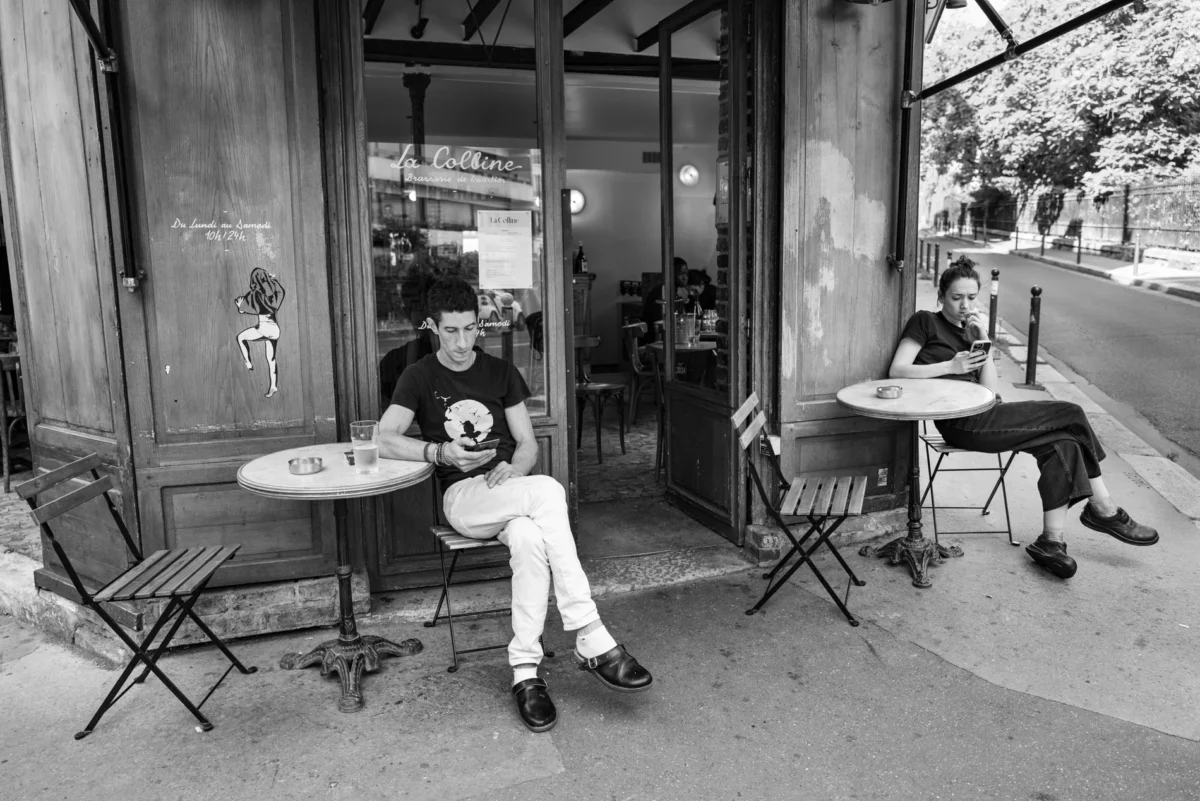 Pascal SC · 02/08/2024 · 1900 x 1268 Cette photographie en noir et blanc montre deux hommes assis à des tables de café en extérieur. Le sujet principal est l’un des hommes, jouant de la guitare, tandis que l’autre est assis à côté d’une porte entrouverte.La composition est symétrique et équilibrée, avec un cadrage serré sur les deux personnages. La lumière semble naturelle, douce et légèrement tamisée, créant des contrastes modérés. L’ambiance est calme et détendue, évoquant une pause conviviale en ville. Le style est documentaire et intimiste, capturant un moment de vie quotidienne.