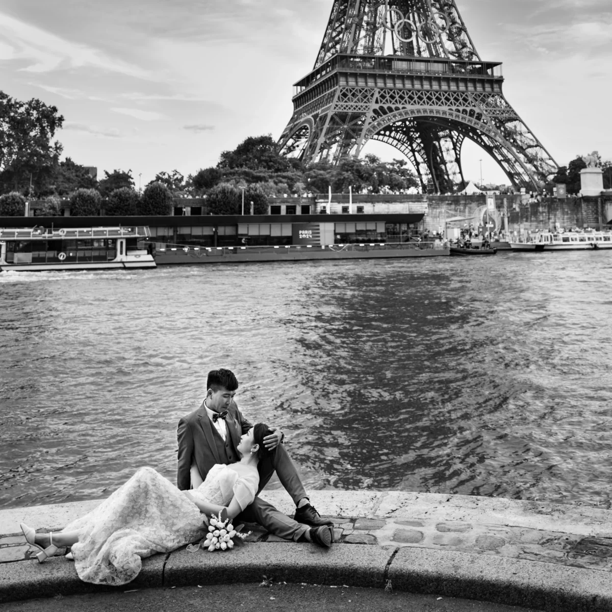 Pascal SC · 07/08/2024 · 1900 x 1900 Cette photographie en noir et blanc met en scène un couple assis sur une berge, le sujet principal étant l’homme en costume et la femme en robe de mariée. La composition joue sur la proximité du couple avec la Seine et l’Eiffel Tour en arrière-plan, créant une ambiance romantique. Le cadrage inclut une partie du paysage urbain parisien, avec des bateaux-mouches sur l’eau. La lumière douce et diffuse accentue les contrastes, renforçant l’émotion. Les tons gris dominent, typiques du noir et blanc, soulignant les textures et les détails. Le style est documentaire et poétique, évoquant une ambiance intime et mélancolique.