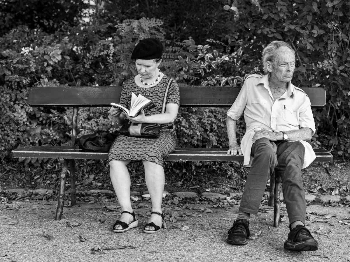 Pascal SC · 01/08/2024 · 1900 x 1425 Cette photographie en noir et blanc met en scène deux personnes âgées assises sur un banc dans un parc. La femme, à gauche, lit un livre ou une lettre, tandis que l’homme, à droite, observe devant lui. La composition est simple et symétrique, avec un cadrage serré sur les sujets.La lumière douce et naturelle crée des contrastes subtils. L’ambiance est calme et introspective, évoquant la sérénité et la tranquillité. Le style est documentaire et réaliste, renforcé par l’absence de couleurs, mettant l’accent sur les détails et les expressions.