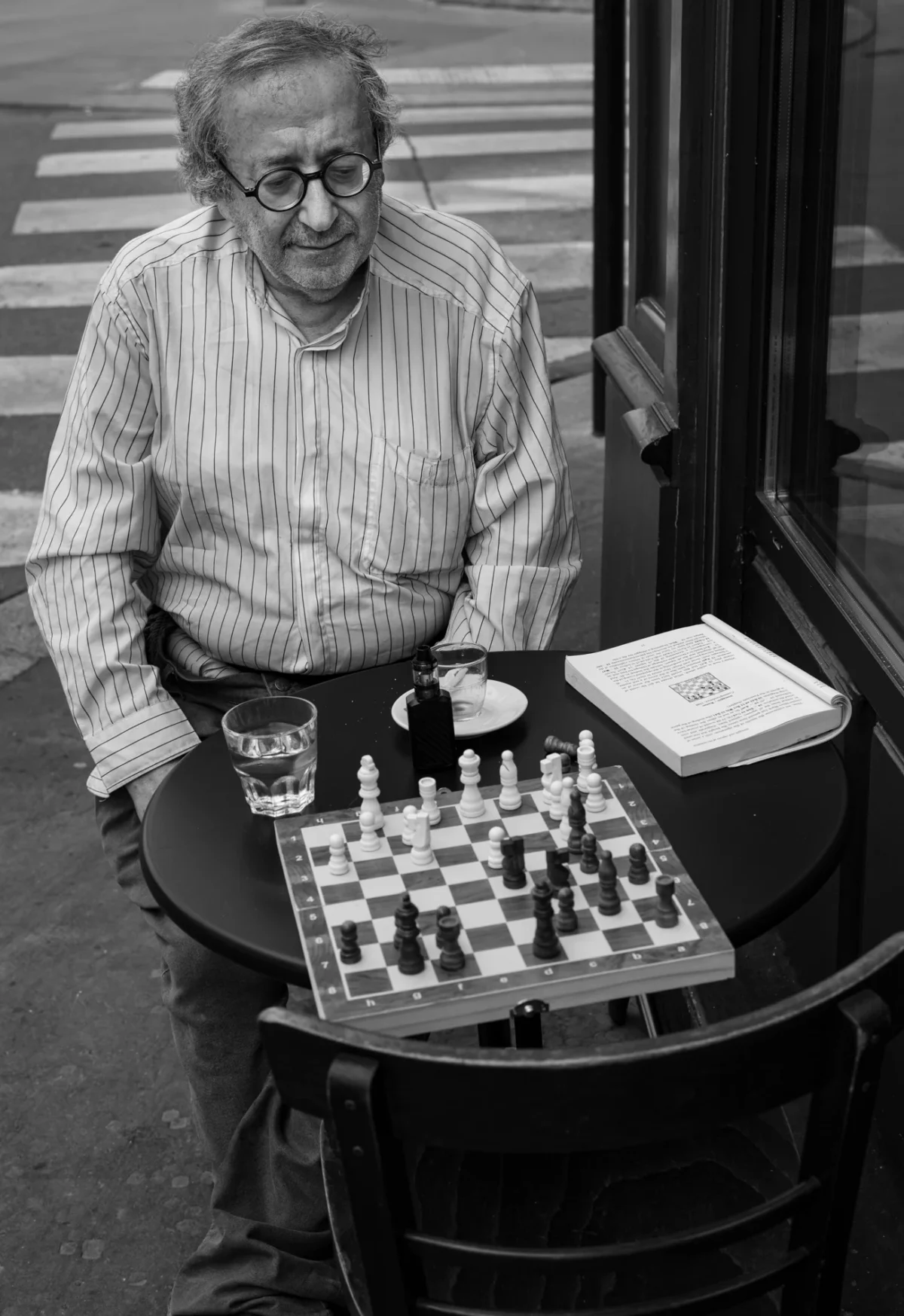 Pascal SC · 01/08/2024 · 1305 x 1900 Cette photographie en noir et blanc met en scène un vieil homme assis, jouant aux échecs. Le sujet principal est cet homme, concentré sur la partie.La composition montre un cadrage serré sur la table à échecs et le joueur, avec un arrière-plan flou de trottoir et de rue. La lumière semble naturelle, douce et légèrement rasante, créant des contrastes modérés. Les tons dominants sont les gris et noirs, typiques du noir et blanc, avec des détails en clair-obscur.Le style est documentaire et intime, évoquant une ambiance calme et contemplative, presque mélancolique.