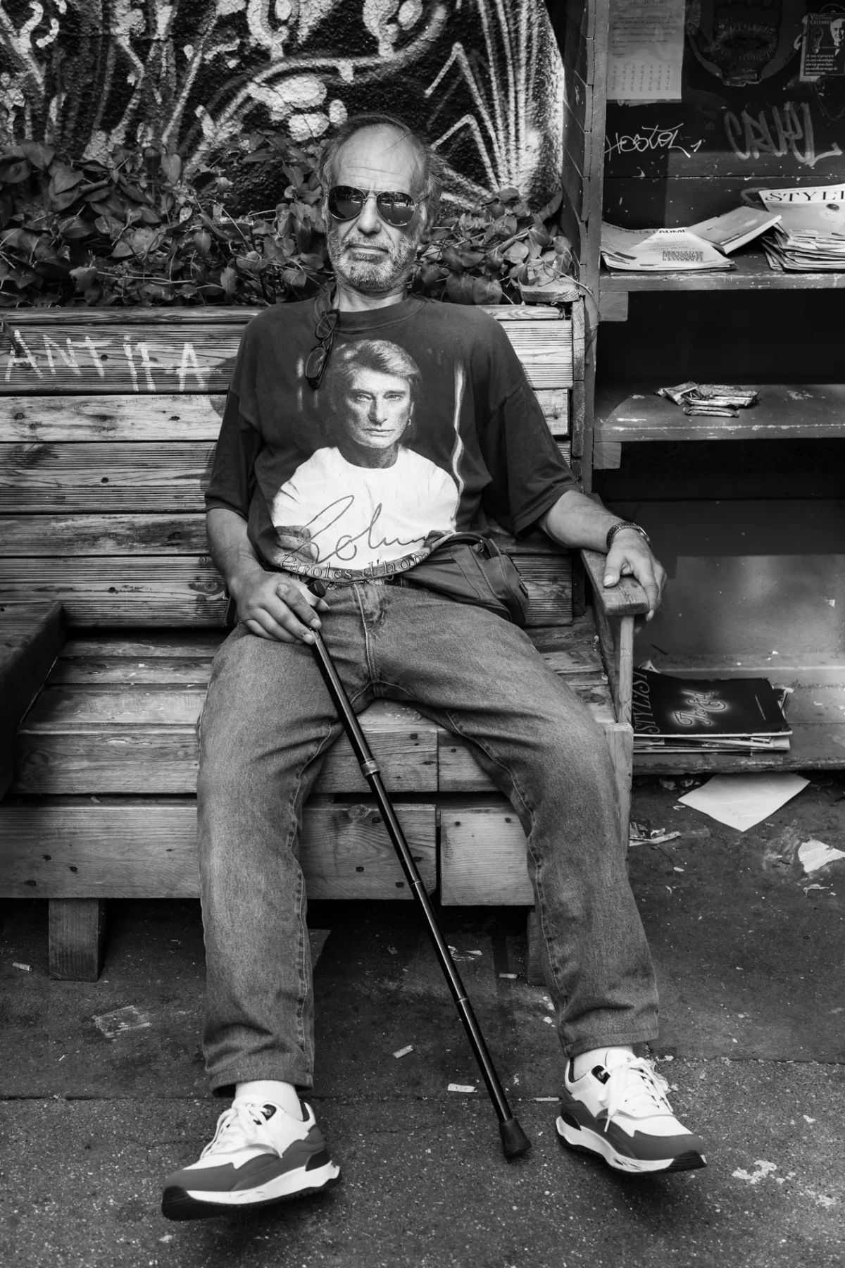 Pascal SC · 01/08/2024 · 1267 x 1900 Cette photographie en noir et blanc met en scène un homme âgé, sujet principal, assis sur un banc avec une canne, tenant un enfant. La composition est simple et centrée, avec un cadrage serré sur les deux personnages. L’arrière-plan, légèrement flou, montre des éléments urbains et des plantes.La lumière semble naturelle, douce et légèrement contrastée. Les couleurs dominantes, bien que monochromes, varient des tons clairs aux ombres profondes, accentuant les détails.Le style est documentaire et réaliste, évoquant une ambiance calme et intime, presque mélancolique.