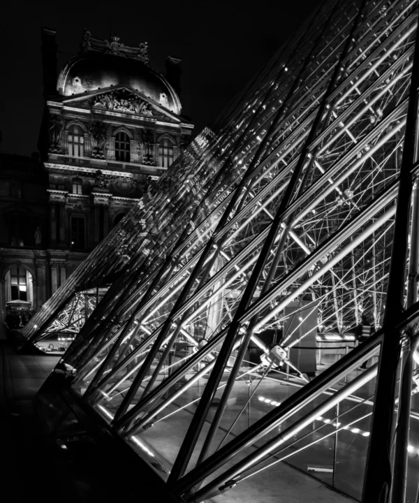 Pascal SC · 10/01/2026 · 1601 x 1920 Cette photographie en noir et blanc met en scène une structure métallique géométrique complexe, probablement une sculpture ou une installation artistique. Le sujet principal est cette structure transparente et entrelacée, reflétant un bâtiment historique en arrière-plan.Le cadrage est serré, centré sur les lignes diagonales de la structure, créant un jeu de perspectives et de profondeur. La lumière, bien que tamisée, accentue les contours et les reflets, renforçant l’aspect architectural et artistique. L’ambiance est à la fois moderne et mystérieuse, mêlant rigueur géométrique et élégance classique. Le style est épuré et minimaliste, typique de la photographie artistique contemporaine.