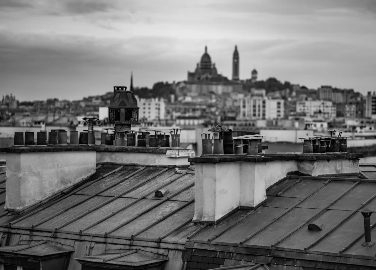 Pascal SC · 19/10/2025 · 1920 x 1379 Cette photographie en noir et blanc montre une ville avec pour sujet principal une vue panoramique de toits et d’un dôme lointain, probablement une cathédrale. La composition met en avant une perspective descendante depuis les toits vers l’horizon.Le cadrage est large, capturant une étendue urbaine dense. La lumière est douce et diffuse, typique d’un jour nuageux, créant des contrastes atténués. Les tons dominants sont les gris et noirs, renforçant une ambiance mélancolique et intemporelle. Le style est documentaire et sobre, évoquant une atmosphère calme et nostalgique.